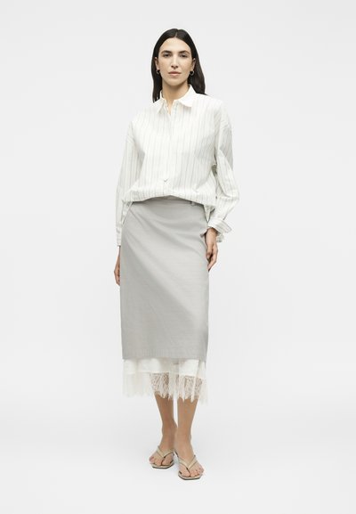 Γυναίκα που στέκεται φορώντας λευκό πουκάμισο με λεπτές ρίγες, ανοιχτογκρι midi φούστα με λευκή δαντέλα στη βάση και μπεζ ανοιχτά πέδιλα σε λευκό φόντο.