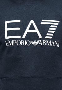 A tengerészkék pulóver nagy fehér "EA7" és "Emporio Armani" felirattal, valamint egy stilizált sas logóval díszített, puha, texturált anyagból készült.