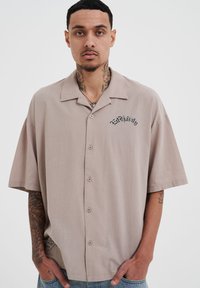 Beige overhemd met korte mouwen en een relaxte snit, met een knoopsluiting aan de voorkant en een zwart geborduurd logo aan de linkerkant. Zachte stoftextuur.