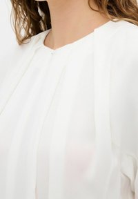 Femme portant un chemisier blanc avec des détails plissés près de l'encolure et des cheveux bruns ondulés visibles sur un fond clair.