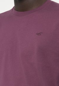 Tricou din bumbac violet, cu guler rotund, mâneci scurte și un mic logo brodat pe partea stângă a pieptului. Textură netedă și design casual.