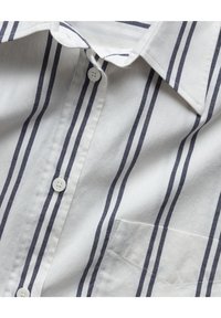Chemise blanche avec des rayures noires verticales, des boutons blancs, une poche avant et un col pointu, vue en gros plan.