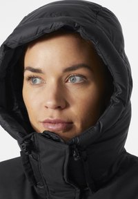 La chaqueta aislante negra presenta un cuello alto y una capucha con cordón. Textura suave con detalles cosidos cerca de la cremallera y la zona del mentón.