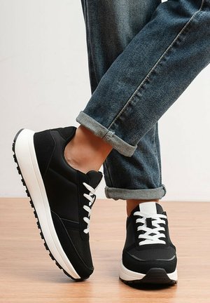 Personne portant des baskets noires et blanches avec des lacets blancs et un jean bleu marine retroussé, debout sur un sol en bois.