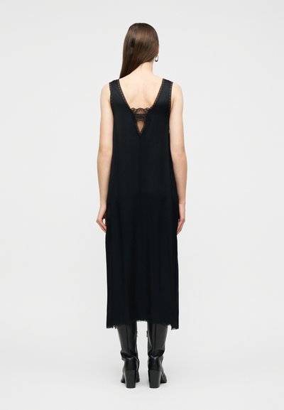 Γυναίκα με μαύρο midi φόρεμα χωρίς μανίκια με δαντελένια λεπτομέρεια στην πλάτη, φοράει μαύρα μπότες με τακούνι, στέκεται μπροστά σε έναν απλό φωτεινό φόντο.