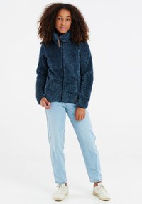 Giacca in pile di colore blu scuro, con zip e colletto alto, caratterizzata da una superficie testurizzata. Abbinata a jeans azzurri chiari e sneakers bianche.