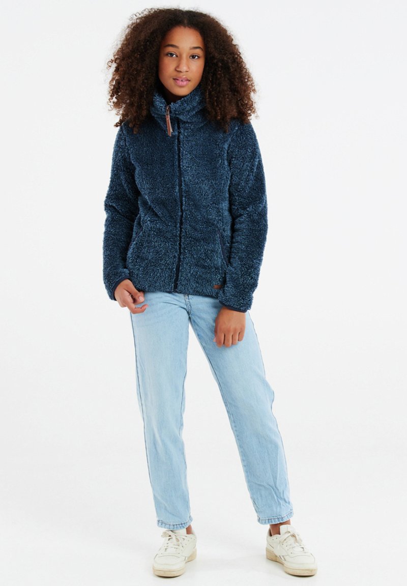 Giacca in pile di colore blu scuro, con zip e colletto alto, caratterizzata da una superficie testurizzata. Abbinata a jeans azzurri chiari e sneakers bianche.