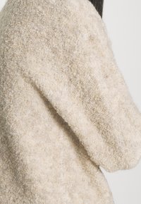 Béžový svetr s texturou, volného střihu a širokými rukávy, vyrobený z jemného, huňatého materiálu. Minimalistický design bez viditelných zapínání.