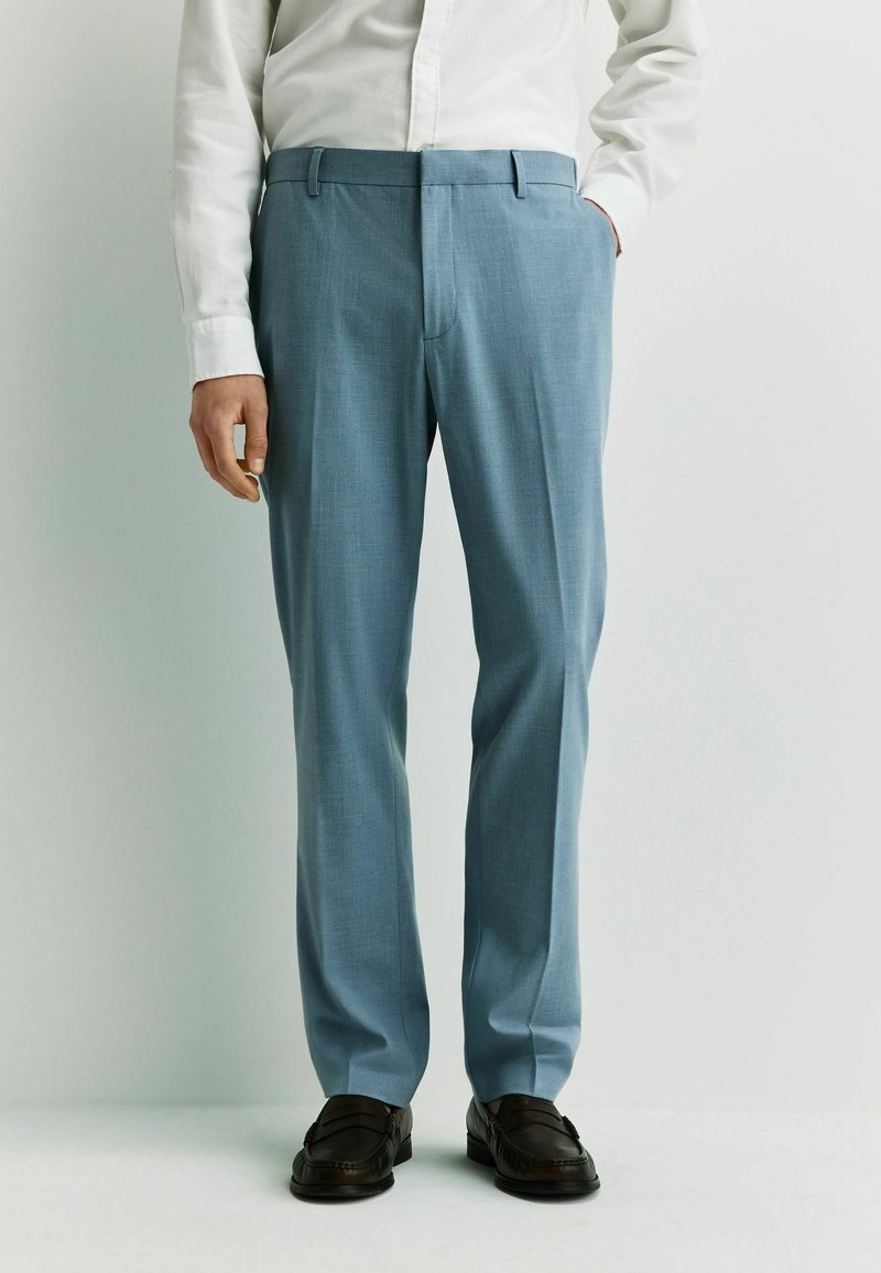 Homme portant un pantalon bleu clair sur mesure, une chemise blanche rentrée, des mocassins en cuir noir, debout avec une main dans la poche.