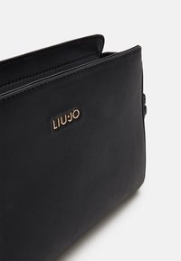 Sorte læderhåndtaske med glat tekstur, der har et guldlogo "LIU JO" på forsiden. Designet inkluderer en topåbning og minimalt hardware.