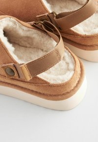 Barna nubuk slip-on lábbeli, puha belső béléssel, széles textil pánttal és bézs gumitalppal.
