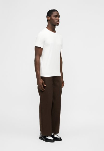 Άνδρας που στέκεται φορώντας λευκό κοντομάνικο μπλουζάκι, καφέ παντελόνι με φαρδύ πόδι και μαύρα και λευκά παπούτσια φορεσιάς μπροστά σε απλό φόντο.