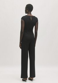 Schwarzer, gerippter Jumpsuit mit einem Schlüssel-Ausschnitt auf der Rückseite, kurzen Ärmeln und weitem Bein, aus weichem, dehnbarem Stoff. Minimalistisches Design.