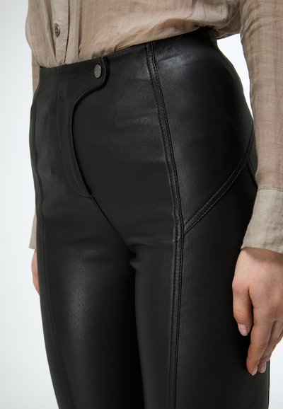 Close-up van een persoon die zwarte faux lederen broek met hoge taille draagt met een knoop aan de voorkant en een beige shirt met lange mouwen, hand rustend langs de zijkant.