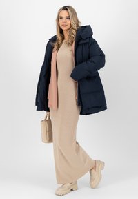 Giacca puffer blu navy con cappuccio, vestito maxi beige a coste, sciarpa rosa chiara, borsa a spalla beige e scarpe beige con suola spessa.