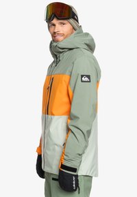 Chaqueta de nieve en bloques de color verde y naranja con capucha, bolsillos con cremallera y parche logo, combinada con manoplas negras y gafas.