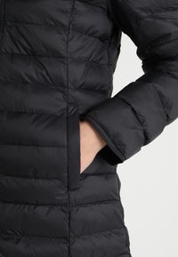 Chaqueta acolchada negra con un patrón de rombos, que presenta un bolsillo lateral y una manga con puño. La tela parece brillante y ligera.