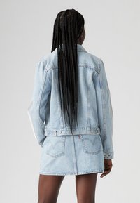 Giacca di jeans azzurro chiaro con due tasche posteriori, hardware argentato e colletto alto. La giacca è abbinata a una gonna di jeans coordinata.