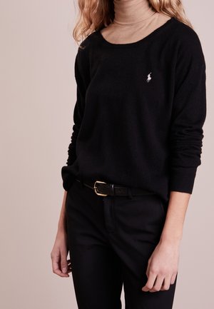 Femme portant un pull noir avec un logo brodé blanc, un col roulé beige, un pantalon noir et une ceinture noire avec une boucle dorée sur un fond neutre.
