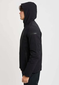 Giubbotto softshell nero con cappuccio aderente, tasche laterali con zip e una finitura texturizzata. Presenta un patch con logo sul maniche.