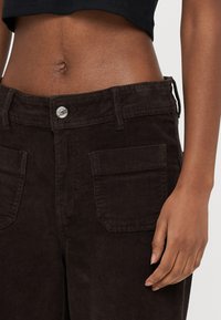 Calças de bombazina castanha com cintura alta, apresentando um fecho de botão metálico e dois bolsos frontais. A textura é canelada, criando um padrão distinto.