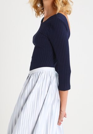 Kobieta ubrana w granatowy prążkowany top z odkrytymi ramionami i rękawami do połowy przedramienia oraz wysoką, białą plisowaną spódnicę.