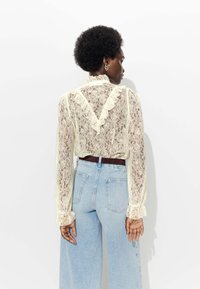 Blouse en dentelle crème avec un décolleté en V et des accents volantés. Manches longues transparents et coupe décontractée, associée à un jean large bleu clair.