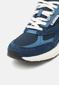 Baskets Tommy Jeans bleues avec lacets blancs, panneaux en maille et en daim, et semelle en caoutchouc blanche, montrant les détails avant et latéraux.
