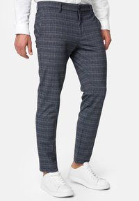 Mann in schmal geschnittenen blauen und grauen karonierten Hosen, die mit weißen Sneakers und einem weißen Langarmhemd kombiniert sind, steht vor einem weißen Hintergrund.