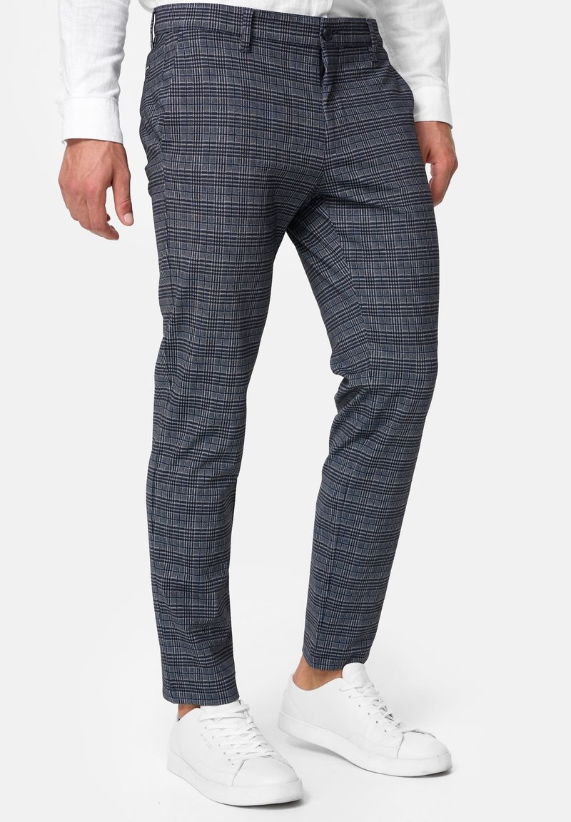 Mann in schmal geschnittenen blauen und grauen karonierten Hosen, die mit weißen Sneakers und einem weißen Langarmhemd kombiniert sind, steht vor einem weißen Hintergrund.