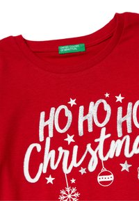 Rotes Baumwoll-T-Shirt mit weißem Glitzertext "HO HO HO Weihnachten" sowie Schneeflocken und Sternen. Das Etikett trägt die Aufschrift "United Colors of Benetton."