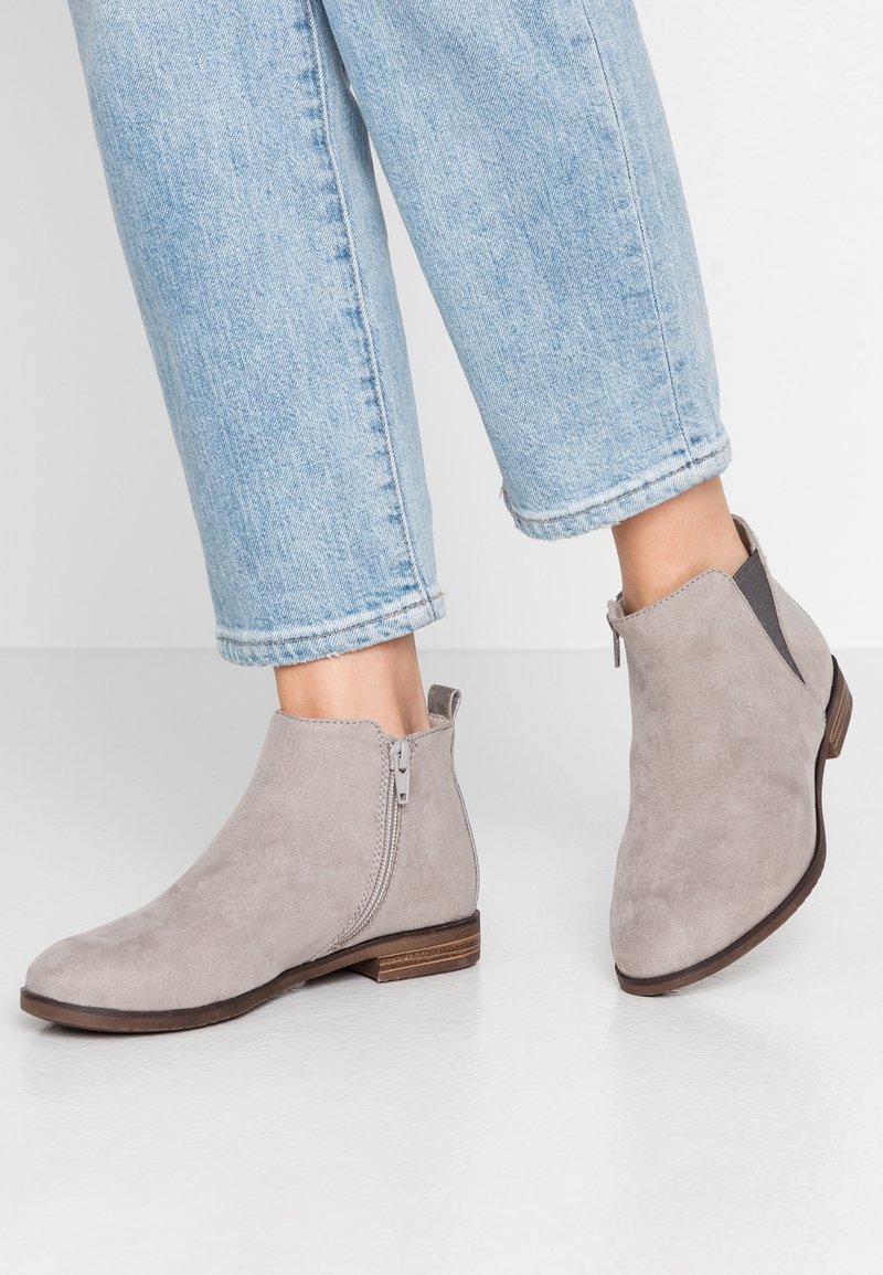 Personne portant un jean court bleu clair et des bottines en suède gris clair avec des fermetures éclair latérales et des petits talons en bois.