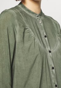 Camicia verde con bottone, realizzata in tessuto testurizzato, dotata di colletto alla mandarina, leggere arricciature e bottoni neri lungo la filettatura frontale.