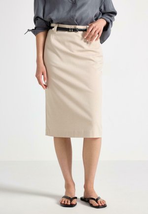 Donna che indossa una gonna beige a tubino al ginocchio con cintura nera, camicia grigia con maniche arrotolate e sandali neri con punta aperta, in piedi davanti a sfondo bianco.