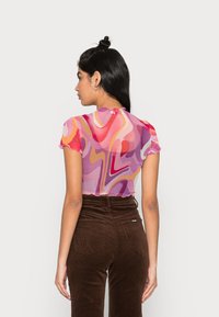 Top de mangas curtas, transparente, com um padrão ondulado em rosa e roxo, com bordas rendadas, combinado com calças de veludo cotelê castanhas de cintura alta com bolsos.