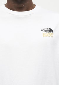 Κοντινό πλάνο ενός ατόμου που φοράει λευκό μπλουζάκι με το λογότυπο "THE NORTH FACE 1966" σε μαύρο και μπεζ χρώμα στο αριστερό μέρος του στήθους.