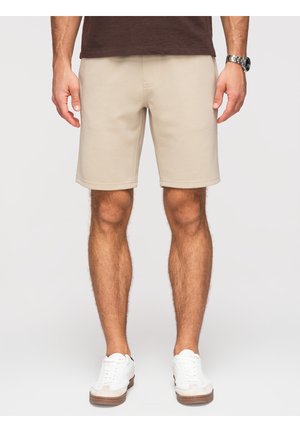 Homme portant un short beige jusqu'aux genoux, des baskets blanches et une chemise marron, debout devant un fond clair uni.