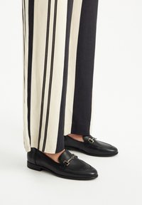 Mocassini in pelle nera con fibbia accentata in oro, abbinati a pantaloni a vita alta a righe in tonalità crema e scure.