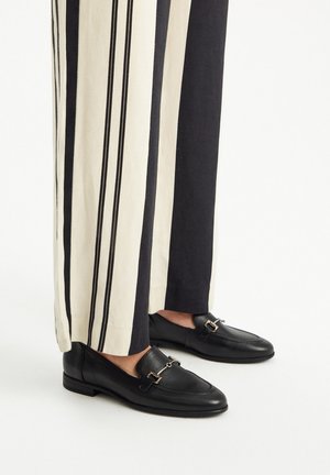 Black leather loafers with a gold-accented buckle, paired with striped high-waisted trousers in cream and dark tones.