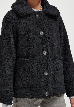 Veste sherpa noire à texture avec boutons, col et poches avant, portée sur un pull gris, associée à un pantalon à carreaux.