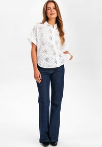 Blusa bianca ricamata con maniche corte, caratterizzata da motivi floreali a intaglio. Abbinata a jeans a zampa d'elefante blu scuro e scarpe nere.
