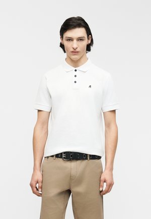 Joven con cabello oscuro, vestido con una camiseta tipo polo blanca, pantalones beige y cinturón negro, de pie frente a un fondo claro y liso.