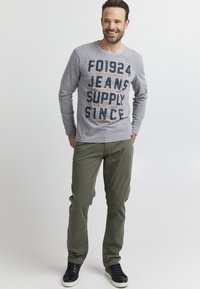Grijze lange mouwen T-shirt met gedurfde meerkleurige tekst, gecombineerd met lichtgroene broek en zwarte sneakers, staand tegen een effen achtergrond.