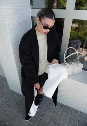 Vrouw die een zwarte jas, een crèmekleurige trui, witte broek en zwarte loafers draagt, zittend op een betegelde rand naast een beige handtas bij een raam.