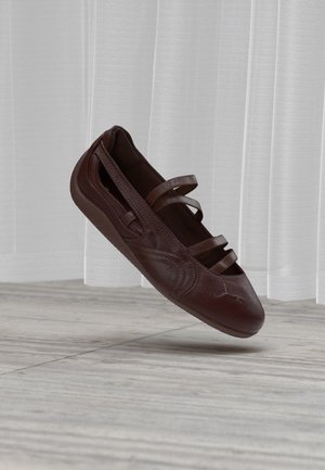 Ballerine en cuir brun foncé avec trois fines lanières sur le dessus, montrée flottant au-dessus d’un sol carrelé clair et de rideaux blancs transparents.