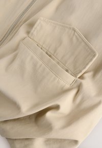 Beige stof met een gladde textuur en fijne ribbels; heeft een gebogen klepzak en een zilveren rits. Ideaal voor casual wear.