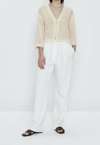 Beige geribde gebreide cardigan met een diepe V-hals en driekwartmouwen, gecombineerd met witte wijde broek en zwarte sandalen.