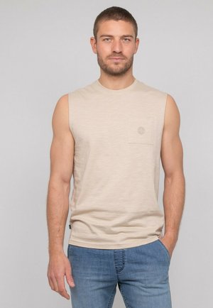 Homme aux cheveux bruns courts et à la barbe, portant un débardeur beige et un jean bleu, debout devant un fond uni gris clair.