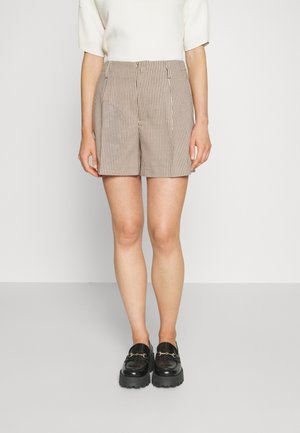 Persona con pantalones cortos a cuadros beige y marrón, camiseta blanca de manga corta y mocasines negros sobre un fondo liso.