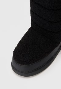 Bota de inverno preta com textura, com parte superior acolchoada e bico e sola reforçados em borracha lisa, sobre um fundo branco.